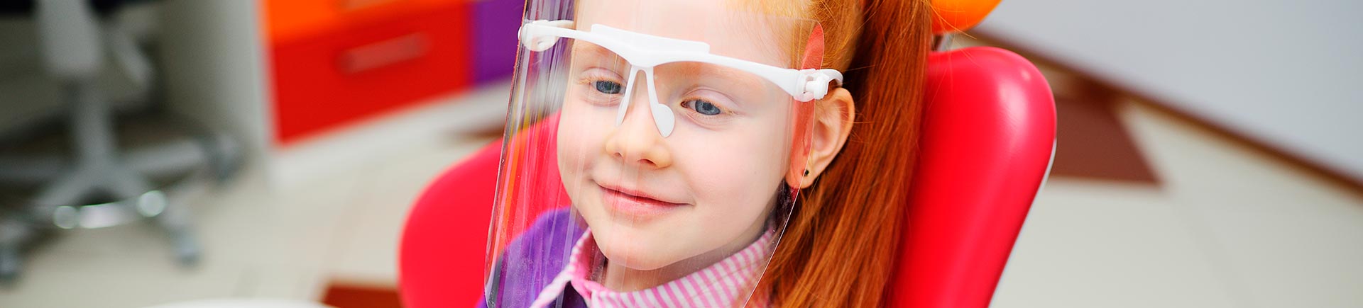 Relaxed little red-haired girl wearing a face shield in a dental chair.