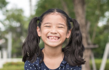 A young girl with black pigtails and a wide smile shows missing front teeth. She is wearing a navy blue polka dot shirt and is outdoors with green trees in the blurred background.